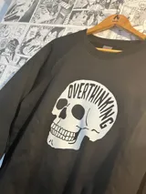 Buzo negro de felpa con cuello redondo y mangas largas. Estampado frontal de una calavera blanca con la palabra "Overthinking".