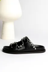 Sandalia tipo mule negra de cuero, con dos tiras ajustables con hebillas metálicas plateadas y apliques de tachas esféricas plateadas. Presenta un borde festoneado en la tira superior y suela de plataforma negra.