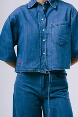 Camisa de denim azul de corte cropped, manga corta ancha, cuello camisero, cierre frontal de botones y un bolsillo aplicado en el pecho.