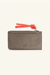 Monedero rectangular de cuero vacuno con textura granulada en color marrón topo. Cuenta con cierre superior metálico y un tirador decorativo de cuero en color naranja con un nudo. Presenta un pequeño logo grabado en el centro.
