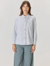 Camisa de algodón color gris claro, con cuello clásico y detalle de volados en la cartera frontal. Presenta mangas largas con puños abotonados y un corte recto.