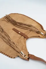 Bolso tipo hobo de gamuza color camel, con flecos largos laterales, correa de hombro de cuero sintético y cierre central con solapa y botón a presión.