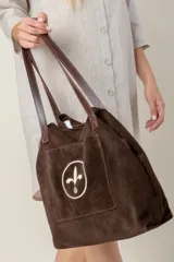 Bolso tipo tote color chocolate, confeccionado en gamuza, con correas largas de cuero marrón. Presenta un bolsillo frontal externo con un diseño bordado circular en color crema.
