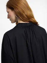 Camisa negra de corte oversize y manga larga con puños abotonados. Presenta un cuello clásico con un lazo fino para anudar en la parte superior y un diseño con bajo asimétrico, siendo más larga en la parte posterior.
