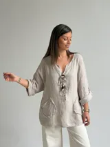 Blusa holgada de lino color beige, con escote en V, mangas tres cuartos con trabilla y bolsillos delanteros aplicados con bordes deshilachados. Cuenta con tiras frontales decorativas con botones de madera.