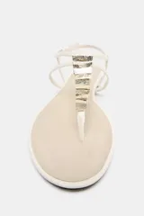 Sandalia plana de color blanco con diseño de tiras finas y separador de dedos. Presenta apliques metálicos decorativos en la tira central y cierre ajustable con hebilla en el tobillo.
