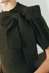 Jersey de punto de manga corta y cuello redondo, en color verde oliva. Presenta un detalle de lazo grande en el frente que le otorga un toque distintivo y femenino.