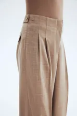 Pantalón sastrero de corte amplio y tiro alto, color beige/tostado con textura sarga. Presenta pinzas frontales y pliegues marcados en las piernas. Tiene cierre con botón y corchete ocultos, y detalles de ajuste con botones forrados en la cintura trasera.