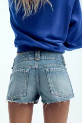Shorts de denim de tiro bajo con cinco bolsillos, detalle de tachas, aplicación de brillos, bajo deshilachado y cierre frontal con cremallera y botones metálicos.