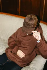 Gorra trucker marrón con frente de tela y parte trasera de rejilla. Presenta un bordado 3D con un diseño abstracto en el frente.