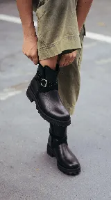 Bota corta de cuero negro con suela track de goma, detalle de tira cruzada con hebilla metálica lateral y paneles de gamuza en contraste.