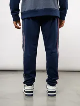 Pantalón deportivo de piqué color azul marino, con franjas laterales en contraste rojo y blanco. Presenta el escudo del Club Nacional de Football bordado en la pierna derecha y una etiqueta conmemorativa en la izquierda.