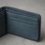 Billetera plegable de eco cuero azul, con múltiples compartimentos para tarjetas, monedero con cierre, tarjetero con visor y dos espacios para billetes. Presenta líneas de pespunte decorativas en la parte superior y el logo de la marca grabado en la esquina inferior.