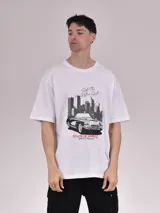 Remera blanca de corte holgado con estampa frontal en tonos negros y rojos que presenta un automóvil clásico frente a un paisaje urbano y la leyenda 'Ride The Historic Route - State of America'.
