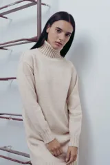 Vestido corto de tejido de punto en color beige, con cuello alto y mangas largas. Presenta puños y ruedo acanalados, con un diseño de calce relajado.