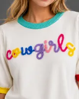 Sweater de punto de manga corta en color blanco, con cuello redondo en color turquesa, puños en colores amarillo y rojo, y borde inferior en color fucsia. Presenta la palabra 'cowgirls' bordada en relieve con letras multicolores.