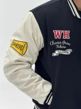 Campera estilo varsity con cuerpo negro y mangas blancas. Presenta parches bordados en el pecho y las mangas, cierre frontal con botones a presión y puños, cuello y ruedo acanalados con rayas blancas y negras.