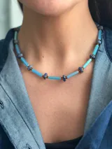 Collar corto de diseño artesanal compuesto por cuentas cilíndricas de vidrio en tono celeste y pequeñas piedras naturales esféricas en tonos azulados, intercaladas con detalles metálicos. Incluye cadena de acero quirúrgico para ajuste de largo.