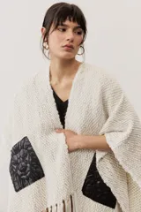 Ruana de punto color beige con detalles geométricos negros y flecos largos tricolores en el borde inferior.
