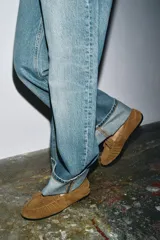 Mocasín de mujer confeccionado en serraje color tostado, con detalle de fruncido y borlas en la parte delantera. Presenta un interior forrado en tejido efecto borreguito y suela plana.