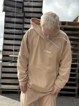 Canguro de tela deportiva color beige, con capucha, cordones ajustables en color blanco y logo bordado en el pecho.