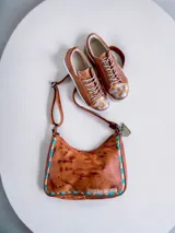 Cartera y bandolera de cuero color marrón tostado con efecto desgastado, detalles de tachas y costuras decorativas en tonos turquesa y marrón, y correa regulable.