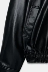 Cazadora negra de efecto piel con cuello subido, manga larga y bolsillos de vivo en el delantero. El bajo está acabado con elásticos y tiene cierre frontal con cremallera metálica oculta por solapa.