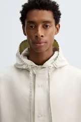 Sobrecamisa de corte oversized confeccionada en tejido compacto de algodón. Presenta cuello subido con capucha ajustable mediante cordones, cierre frontal con botonadura, manga larga con puños abotonados y dos bolsillos de plastrón en la parte frontal.