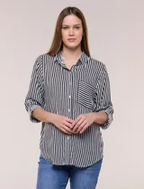 Camisa de corte clásico y silueta relajada con diseño de rayas verticales en blanco y negro. Presenta cuello camisero tradicional, cartera frontal con botones, un bolsillo aplicado en el pecho con rayas horizontales y mangas largas con sistema para arremangar.