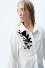 Camisa de corte recto con cuello clásico y cierre frontal de botones. Presenta un parche bordado con mostacillas en tonos blanco y negro en el frente, con diseño de estrellas y formas abstractas.