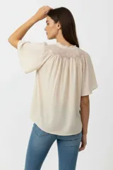 Blusa de viscosa color blanco, con cuello en V y detalle de bordado geométrico en tonos azules en la parte frontal. Presenta mangas cortas con vuelo y un corte holgado.