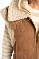 Chaleco estilo gamulán de corte oversize, confeccionado en material tipo gamuza color camel con interior y bordes de corderito sintético color crema. Presenta cierre frontal completo, bolsillos laterales y detalles de tiras decorativas en los hombros.