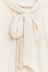 Camisa cruda de manga larga con textura plumeti, cuello redondo y lazada en el mismo tejido. Cuenta con mangas abullonadas terminadas en puños anchos y cierre en la espalda con abertura y botones.