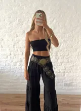 Cinturón ancho de cuero negro con diseño de círculos superpuestos. Presenta apliques metálicos circulares en tono dorado envejecido, algunos lisos y otros con grabado tribal/étnico. Los círculos de cuero tienen ojales metálicos pequeños alrededor de los apliques grandes.