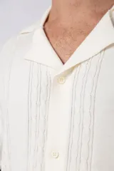 Camisa de punto de manga corta con cuello tipo bowling y cierre frontal de botones. Presenta un diseño texturizado con franjas verticales y detalles de contraste en blanco en los bordes de las mangas.