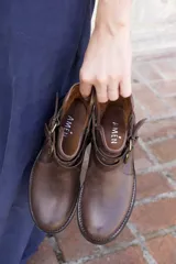 Bota corta de cuero color marrón, con taco bajo cuadrado y punta redondeada. Presenta un diseño estilo western con correas ajustables con hebillas metálicas y herrajes circulares en los laterales.