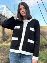 Chaqueta de punto negra con ribetes blancos, cuello redondo, cierre frontal con botones dorados, bolsillos frontales con solapa y puños acanalados.