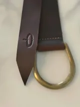 Cinto de cuero flexible diseñado para anudar, con terminación en punta y detalle de marca grabada en el extremo. Incluye una hebilla metálica en forma de D en tono bronce.