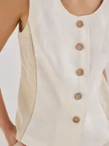 Chaleco sin mangas de lino, con diseño de paneles en color blanco crudo y beige. Presenta escote redondo y cierre frontal con botones de madera marrones.