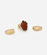 Kit de tres anillos para mujer, elaborados en metal dorado. Incluye un anillo principal con un chaton de cristal geométrico en tono borravino y dos anillos sencillos de superficie pulida.