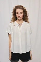 Camisa de manga corta y silueta amplia color blanco, confeccionada en viscosa liviana y de caída fluida. Presenta escote redondo con frunces suaves y abertura frontal con lazo para anudar.