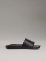 Ojotas tipo slide de color negro con tira superior de piel sintética que presenta un patrón grabado del logo de Calvin Klein. Poseen plantilla ergonómica moldeada y suela de EVA ligera.