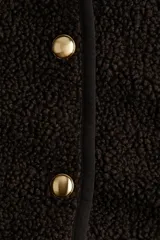 Chaqueta de sherpa suave en color marrón oscuro, con diseño de corte holgado y cuello redondo. Presenta ribetes de cinta finos en los bordes y cierre frontal mediante botones metálicos dorados.