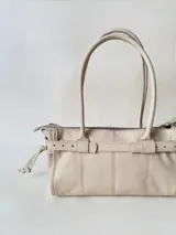 Cartera de cuero de tamaño medio con estructura rígida. Presenta una tira decorativa con tachas y hebillas metálicas en los extremos, además de lazos laterales. Cuenta con doble asa para llevar al hombro y cierre superior.