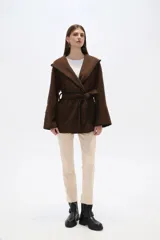 Chaqueta blanca de corte holgado con capucha, cierre frontal cruzado con botones y cinturón de tela ajustable. Presenta detalles de costuras decorativas en color negro en los bordes, solapas, bolsillos y cinturón.