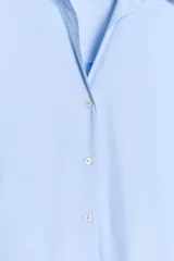 Camisa celeste de popelina, de corte holgado, con cuello solapa, escote en pico y mangas largas. Tiene bajo asimétrico y cierre frontal con botones.