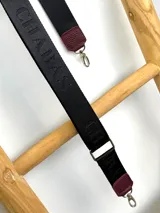 Correa ajustable para cartera confeccionada en cinta tejida con diseño de rayas en negro, blanco y beige, con punteras de cuero sintético negro y herrajes metálicos.