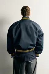 Cazadora bomber estilo universitario, de corte relaxed fit y tejido técnico azul marino, con cuello, puños y dobladillo de rib elástico a rayas amarillas. Cuenta con bolsillos delanteros de plastrón y cierre frontal con botones a presión.