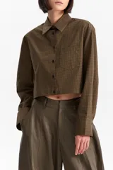 Camisa de manga larga, color verde oliva, con estampado de cuadros pequeños (vichy o grid). Presenta un corte cropped (a la cintura), cuello camisero, botones frontales oscuros y un bolsillo de parche en el pecho con un pequeño bordado.