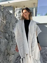 Ruana tejida en lana merino, color beige y crudo, con diseño de trenzas verticales en el frente y terminación con flecos en el ruedo.